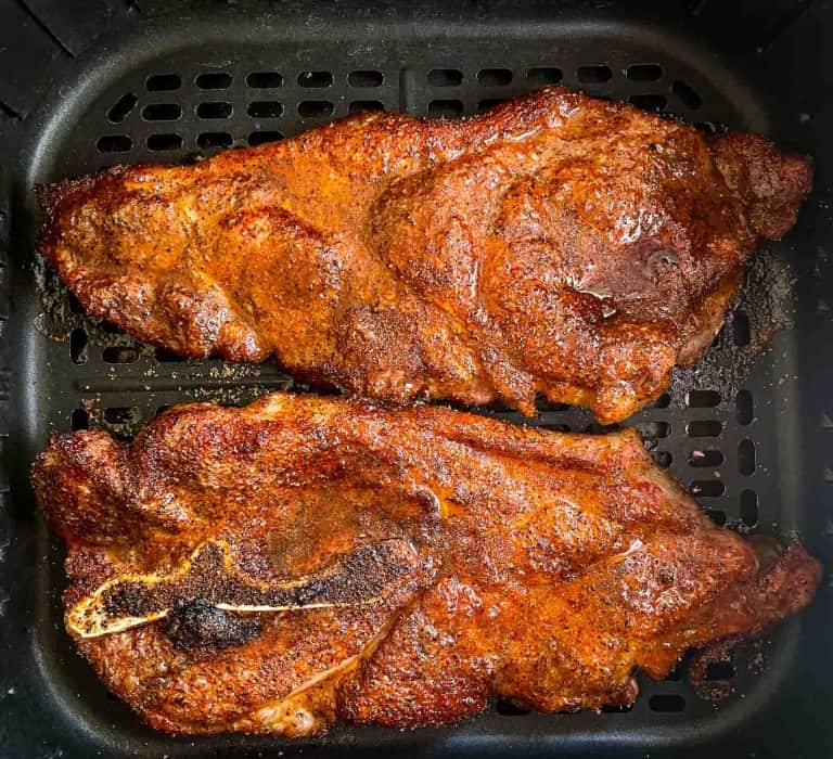 Two seasoned air fryer pork steaks cooking in an air fryer basket, with a visible bone in one of the steaks.