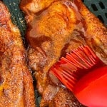 Close-up of barbecue sauce being brushed onto seasoned air fryer pork steak ribs with a red silicone brush on a grill or air fryer tray.
