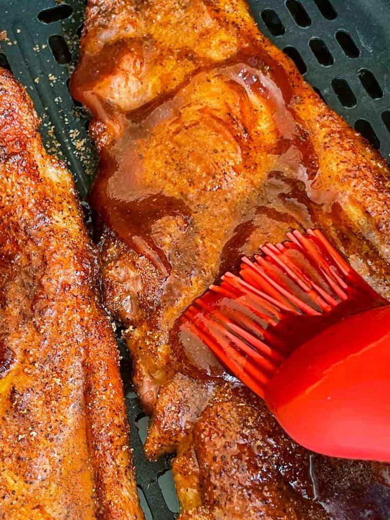 Close-up of barbecue sauce being brushed onto seasoned air fryer pork steak ribs with a red silicone brush on a grill or air fryer tray.