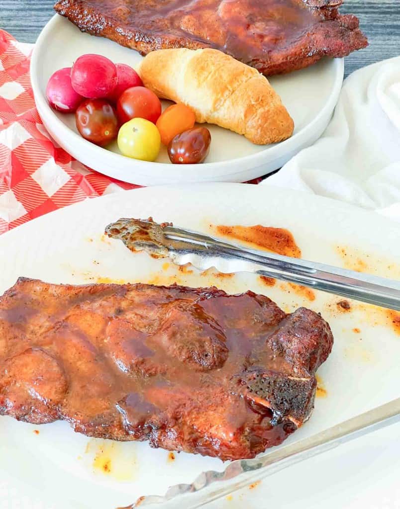 Barbecued steak with sauce on a white plate, metal tongs beside it; in the background, a plate holds an air fryer pork steak, bread roll, and assorted cherry tomatoes.