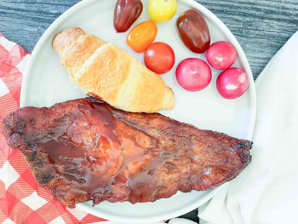A plate with a glazed barbecued rib, an air fryer pork steak, a croissant, three radishes, and four colorful grape tomatoes on a white plate.