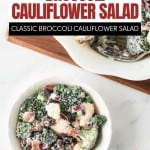 A bowl of creamy Broccoli Cauliflower Salad with chopped red onions, bacon bits, and dressing sits on a white table next to a serving dish.