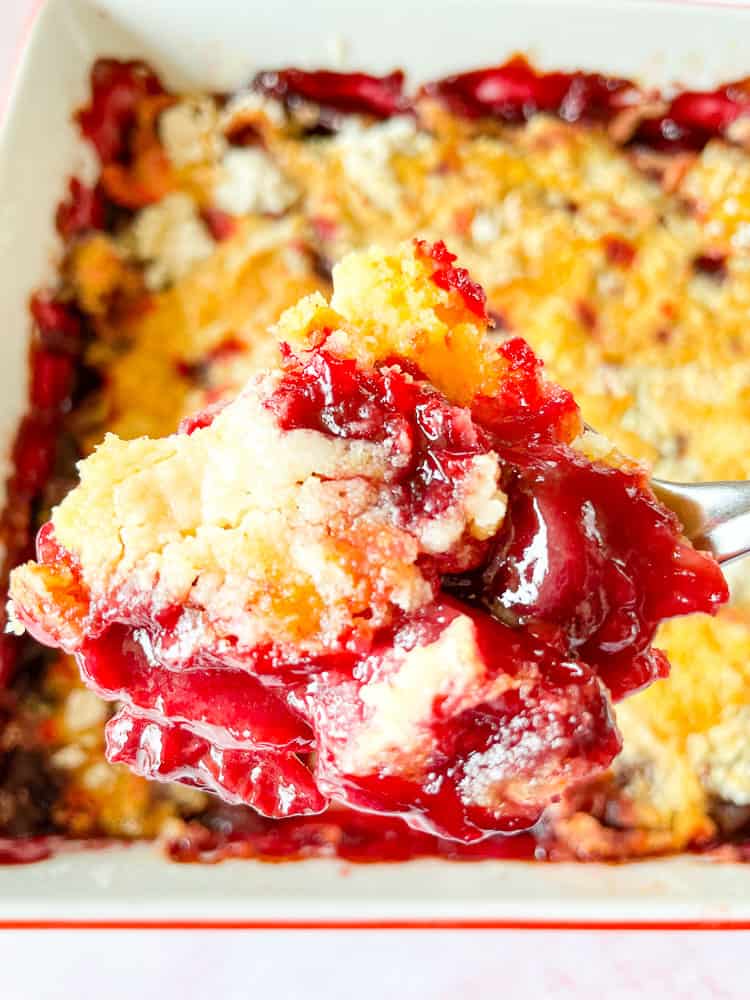 A generous scoop of cherry dump cake held over the pan full of the cake.