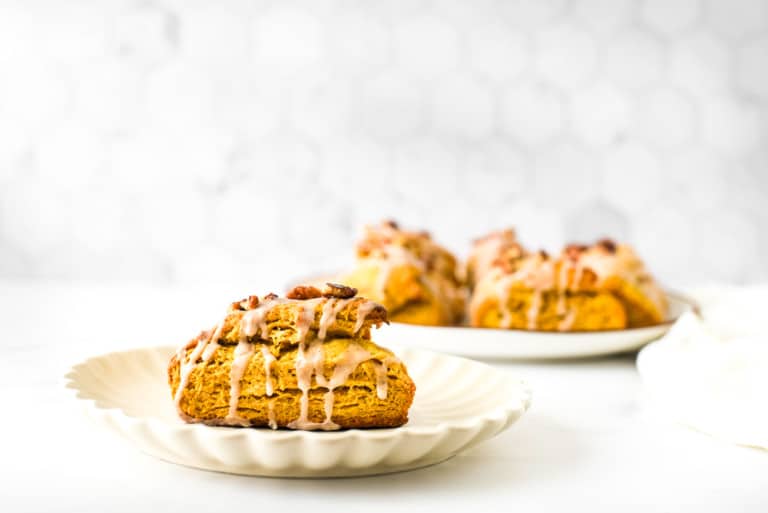A pumpkin scone with cinnamon glaze and candied pecans on a white plate, with more scones in the background.