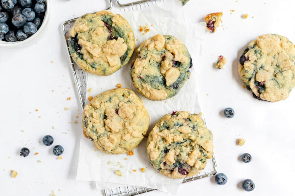 Four blueberry crumb cookies rest on a cooling rack with parchment paper, surrounded by scattered blueberries and cookie crumbs. A bowl of blueberries is partially visible in the upper left corner, adding to the abundance of blueberry delights.