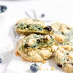 A stack of blueberry cookies sits on white parchment paper. One cookie is broken in half, revealing a soft inside. A bowl of fresh blueberries and scattered crumbs are visible in the background, enhancing the delectable appeal of these blueberry cookies.