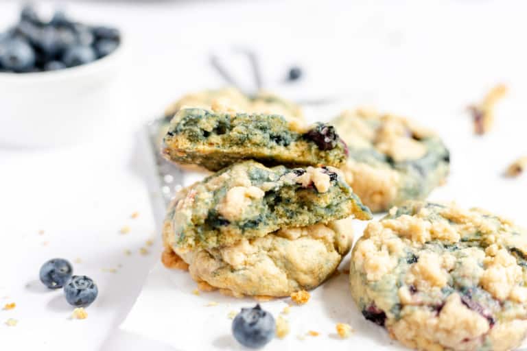 A stack of blueberry cookies sits on white parchment paper. One cookie is broken in half, revealing a soft inside. A bowl of fresh blueberries and scattered crumbs are visible in the background, enhancing the delectable appeal of these blueberry cookies.