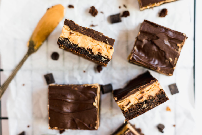 Four chocolate peanut butter brownies are arranged on parchment paper, with a spoonful of peanut butter nearby and scattered crumbs around, reminiscent of Buckeye Brownies.