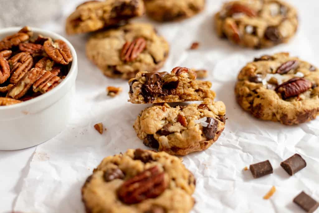 Homemade Cowboy Cookies with pecans scattered on a white surface, some cookies are broken to show a chunky texture.