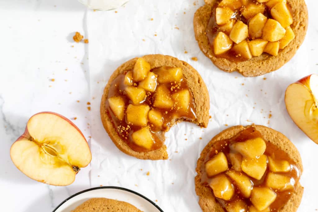 Apple pie cookies on a white background, one with a bite taken out, served alongside fresh apple slices.