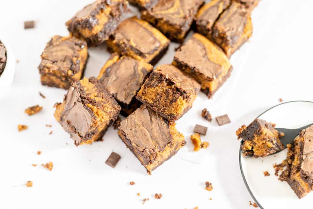 Pumpkin brownies with a fudgy texture, cut into squares and scattered on a white surface, with chocolate chunks and crumbs around them.