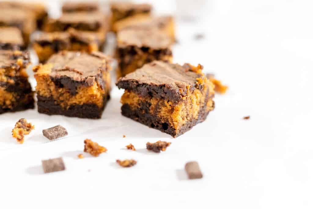 Close-up of pumpkin brownies with a chocolate layer, arranged on a white background with chocolate chunks and small crumbs surrounding them.