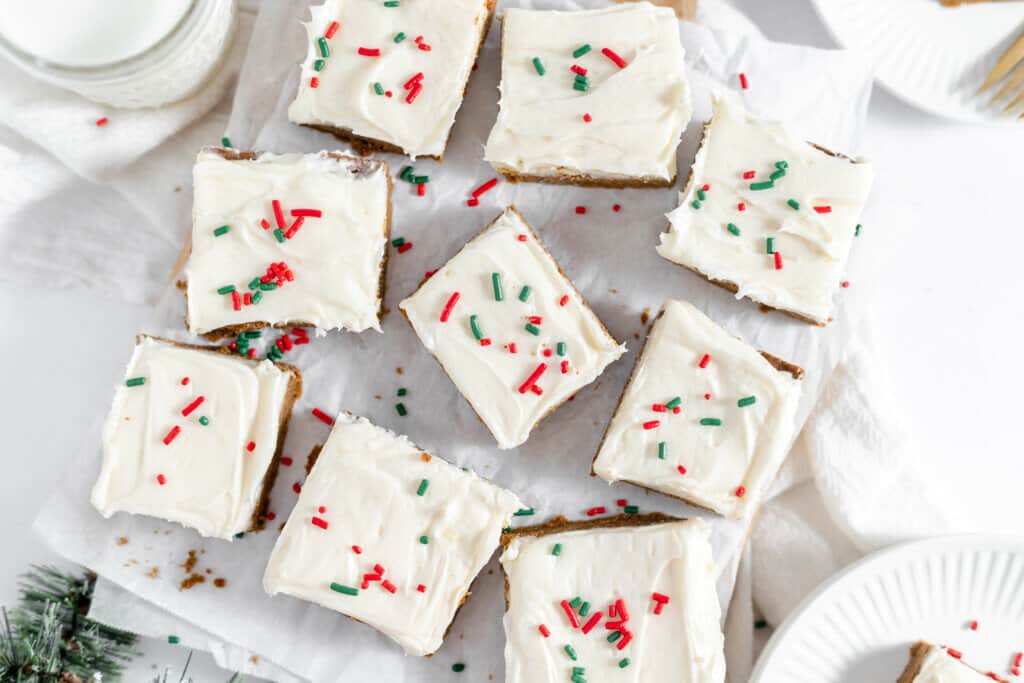 A platter of gingerbread-themed frosted sugar cookie bars decorated with red and green sprinkles, surrounded by pine branches on a white background.
