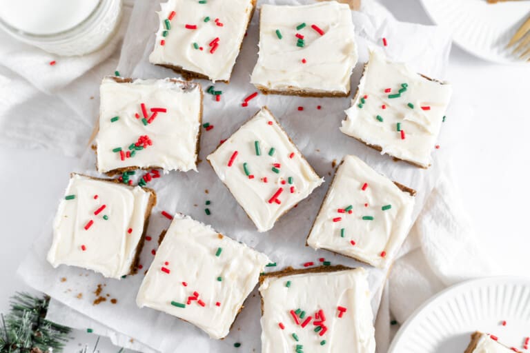 A platter of gingerbread-themed frosted sugar cookie bars decorated with red and green sprinkles, surrounded by pine branches on a white background.