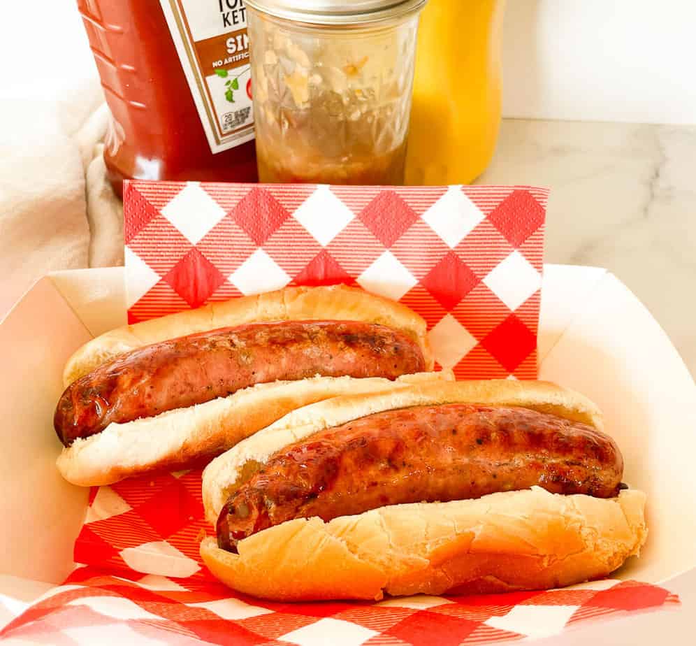 Two grilled Aidells sausages in buns on a red and white checkered paper, with bottles of ketchup and mustard, and a jar of relish in the background.