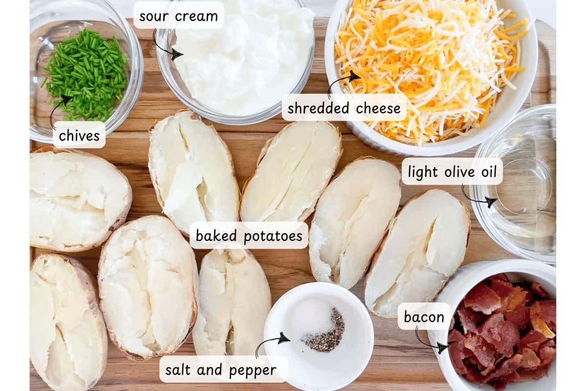 Top-down view of ingredients for loaded baked potatoes, including crispy air fryer potato skins, shredded cheese, sour cream, chives, light olive oil, bacon, and salt and pepper on a wooden board.