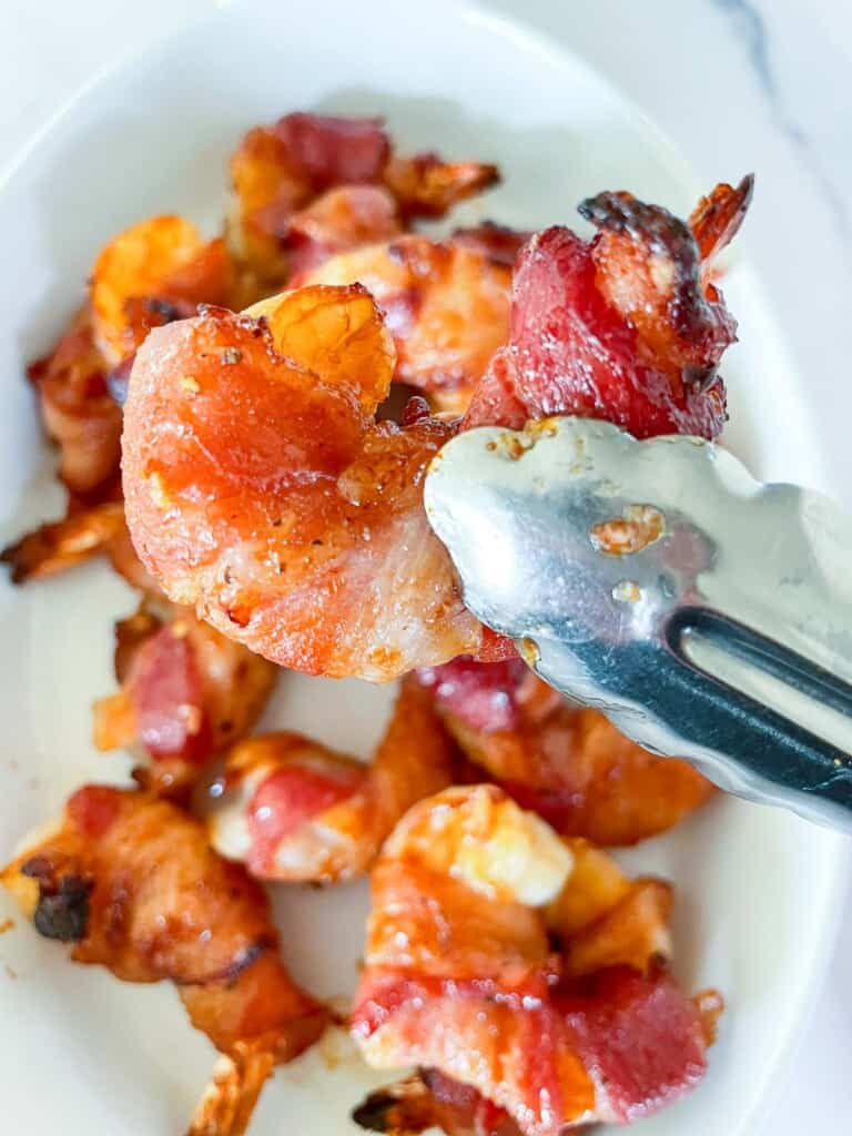 One shrimp in a pair of tongs in front of all the finished bacon wrapped shrimp. 