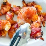One shrimp in a pair of tongs in front of all the finished bacon wrapped shrimp.