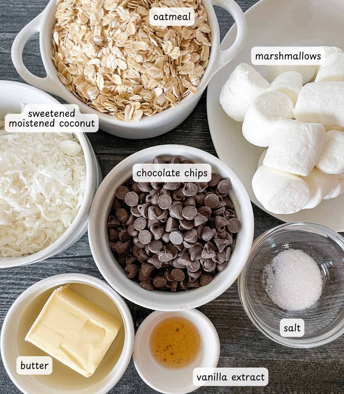 A delightful array of ingredients for Chocolate No Bakes awaits: oatmeal, marshmallows, sweetened coconut, chocolate chips, butter, vanilla extract, and salt are beautifully arranged in bowls on a gray surface.