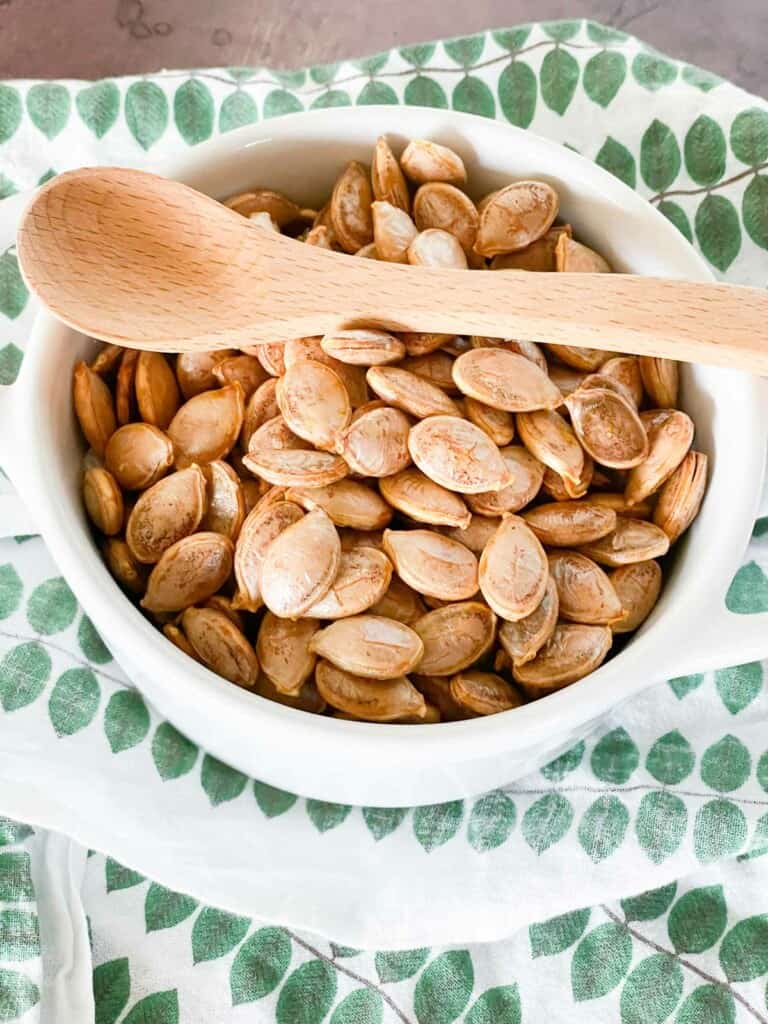 Pumpkin seeds in a white dish with a wooden spoon on a green and white linen.
