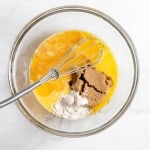 A glass mixing bowl containing a whisked mixture of beaten eggs, brown sugar, and flour for a bourbon pecan pie.