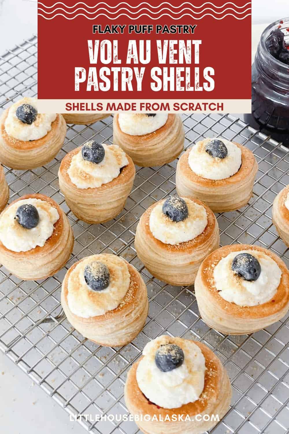 Mini Vol Au Vent puff pastry shells topped with cream and a blueberry, arranged on a cooling rack with a jar of jam nearby.