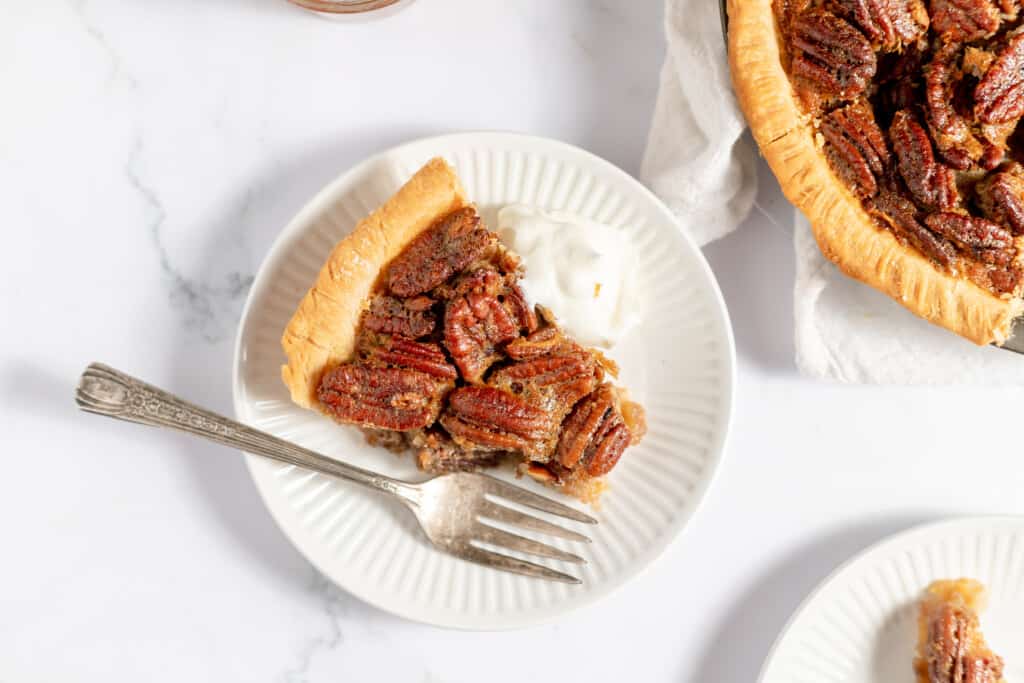 A slice of bourbon pecan pie on a plate with a fork and pecan pie nearby.