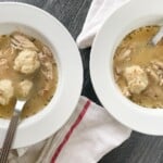 White bowls of chicken and dumplings with spoons and linens.