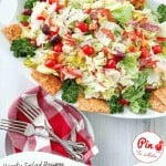 A platter of antipasto salad with lettuce, meats, cheese, olives, tomatoes, and crackers is garnished with parsley. Two forks rest on a red-and-white napkin beside the delicious antipasto salad dish.