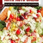 A bowl of antipasto salad with lettuce, cherry tomatoes, olives, cheese, deli meats, and croutons, all topped with fresh herbs and cracked black pepper.