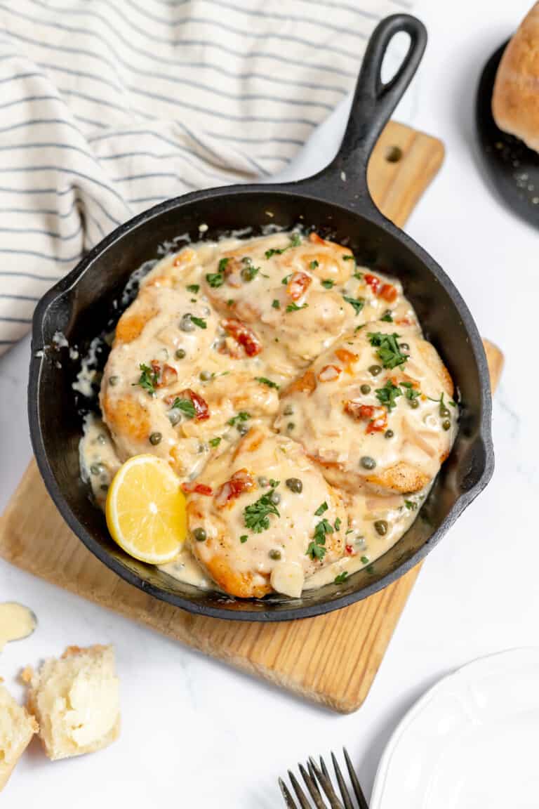 A skillet of creamy chicken piccata with capers and slices of lemon, garnished with parsley, served on a wooden trivet next to a striped cloth and pieces of bread.