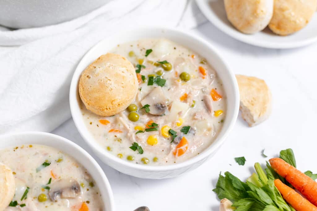 A bowl of creamy Chicken Pot Pie Soup with peas, carrots, and mushrooms, served with a bread roll on a white tablecloth.