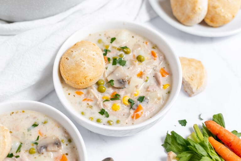 A bowl of creamy Chicken Pot Pie Soup with peas, carrots, and mushrooms, served with a bread roll on a white tablecloth.