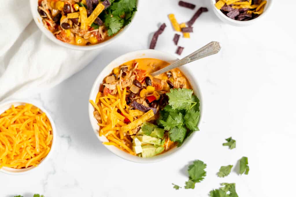 A bowl of chicken taco soup topped with avocado, cheese, cilantro, and tortilla strips, served on a white marble surface.