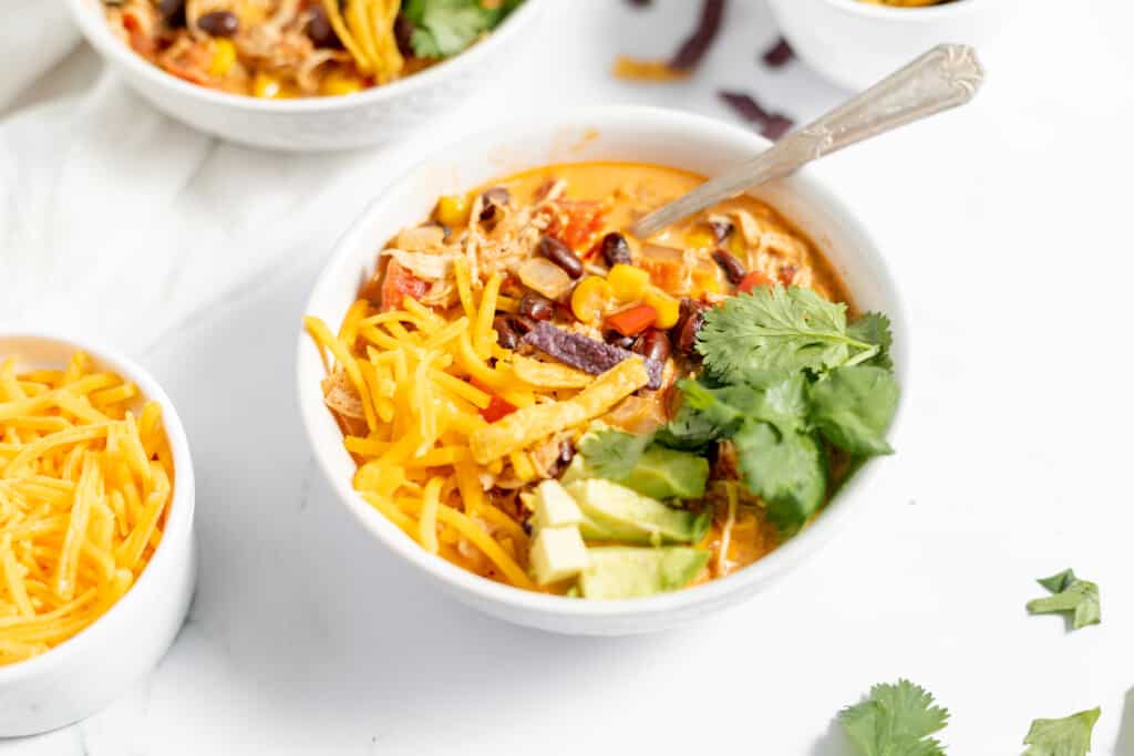 A bowl of chicken taco soup topped with avocado, cheese, cilantro, and tortilla strips, served on a white marble surface.