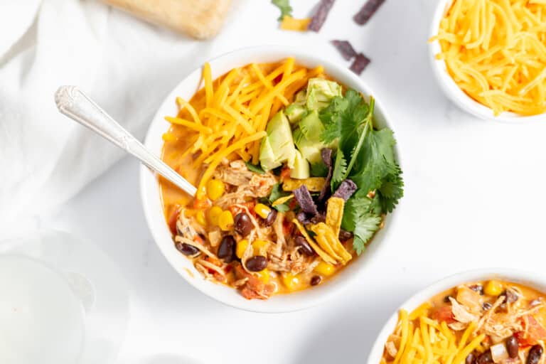 A bowl of chicken taco soup garnished with shredded cheddar cheese, avocado slices, fresh cilantro, and crispy tortilla strips, accompanied by a silver spoon and a small bowl of extra cheese on the side.