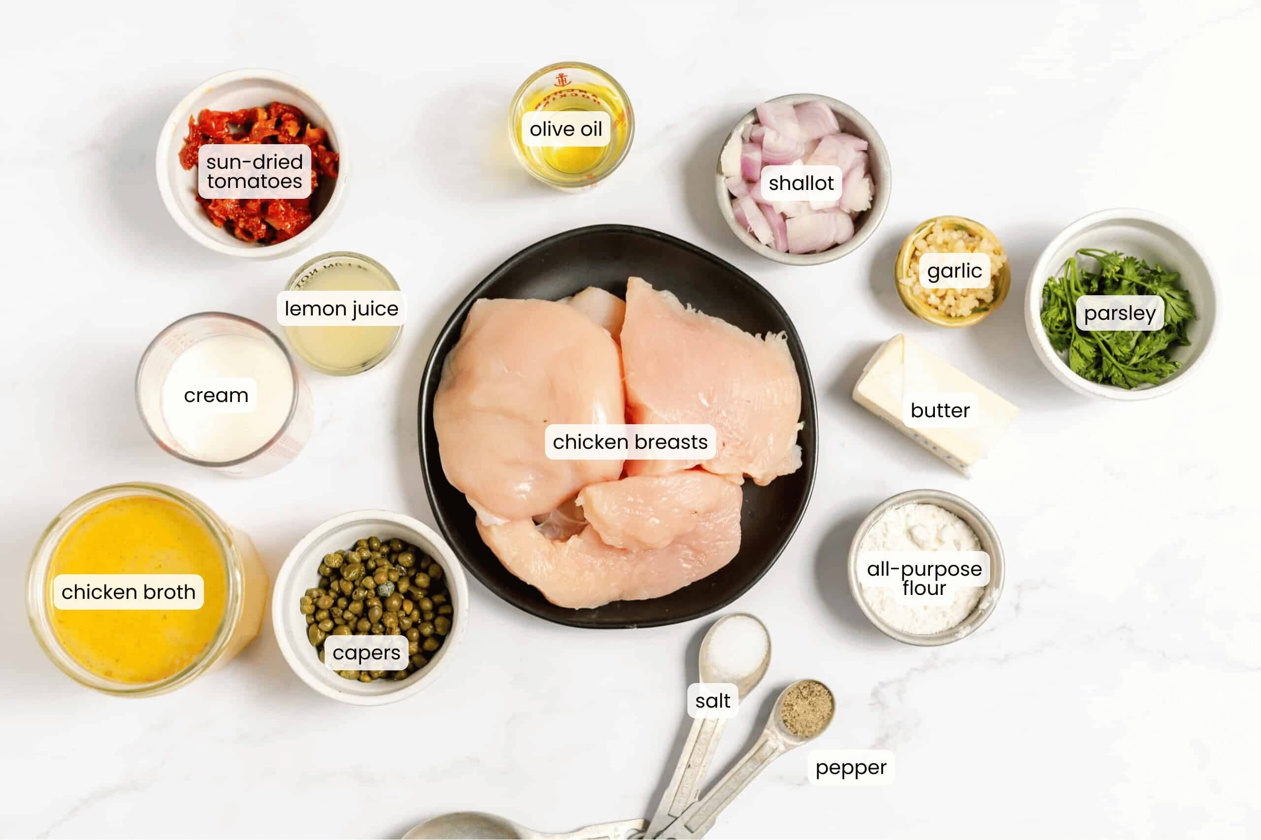 Top-down view of chicken piccata ingredients, including chicken breasts, sun-dried tomatoes, capers, broth, cream, lemon juice, olive oil, shallot, garlic, butter, parsley, flour, salt, and pepper.