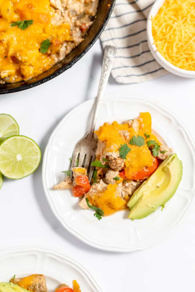 Chicken Fajita Casserole on a white plate with avocado and lime and a skillet of casserole.