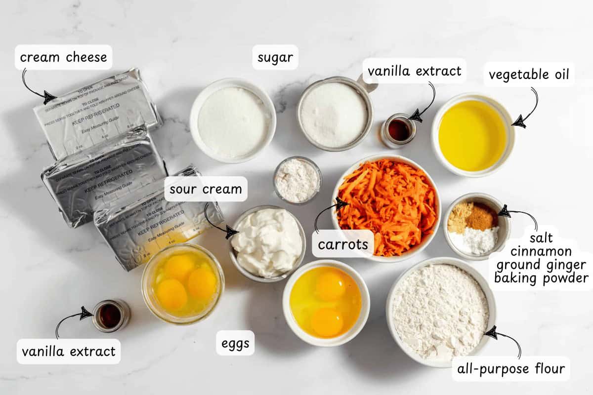 Ingredients for a delightful carrot cake cheesecake, like cream cheese, sour cream, eggs, sugar, vanilla extract, vegetable oil, carrots, spices, salt, baking powder, and all-purpose flour are elegantly arranged on a marble surface.