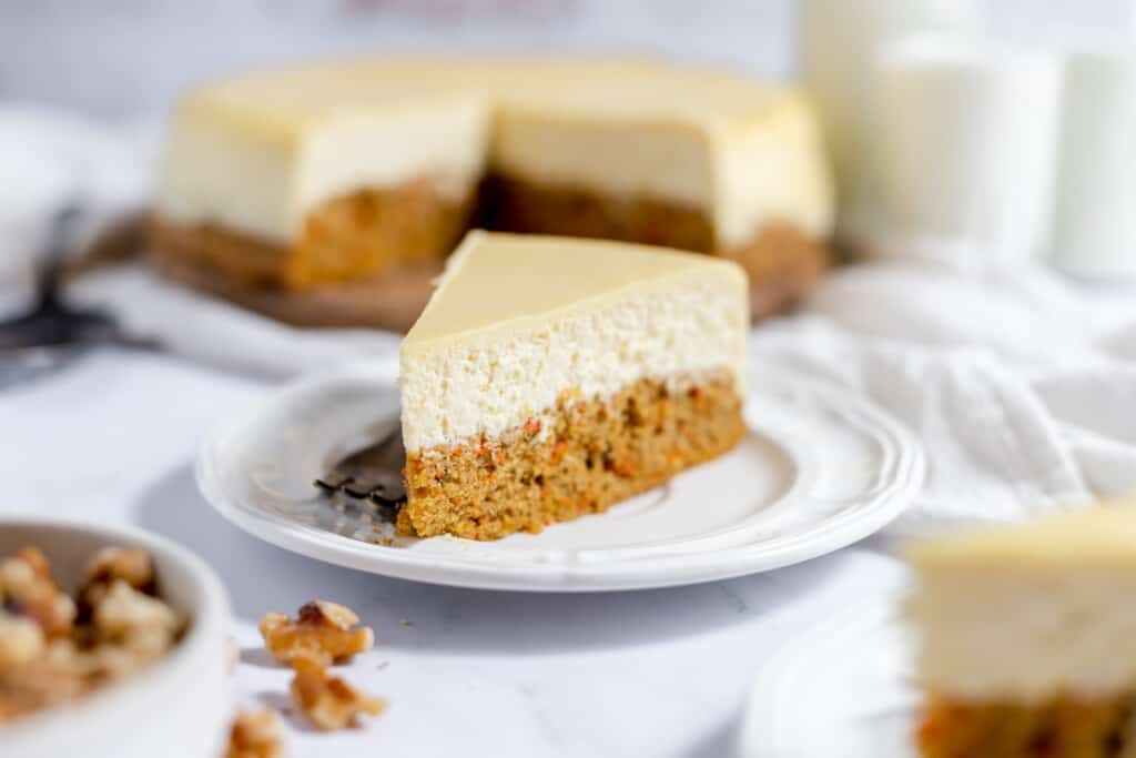 A slice of carrot cake cheesecake on a plate with the cheesecake behind it.