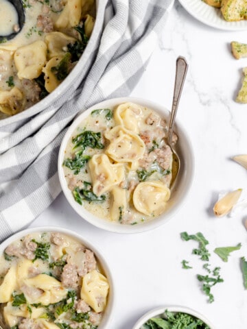 Bowl of creamy tortellini soup with a spoon in it, and other bowls of soup and a dutch oven.