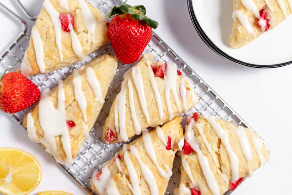Strawberry scones drizzled with lemon glazed on a cooling tray alongside fresh strawberries and lemons.