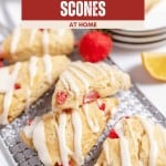 Four strawberry scones with white icing are displayed on a cooling rack, showcasing the irresistible appeal of homemade Strawberry Scones, with lemon slices and fresh strawberries nearby. A stack of plates is in the background.