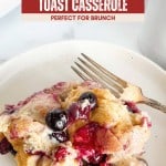 A serving of blueberry French toast casserole sits on a white plate with a fork, the warm blueberry French toast casserole topped with juicy blueberries and a drizzle of syrup.