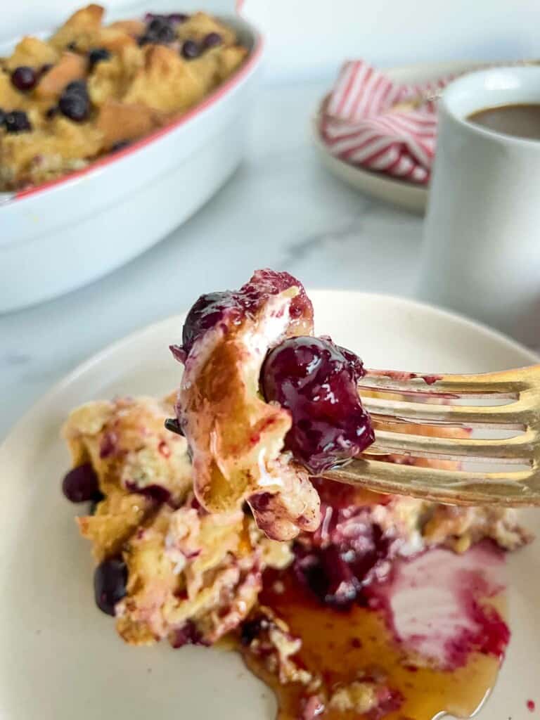 A bite of blueberry casserole with the casserole and coffee cup in the background. 