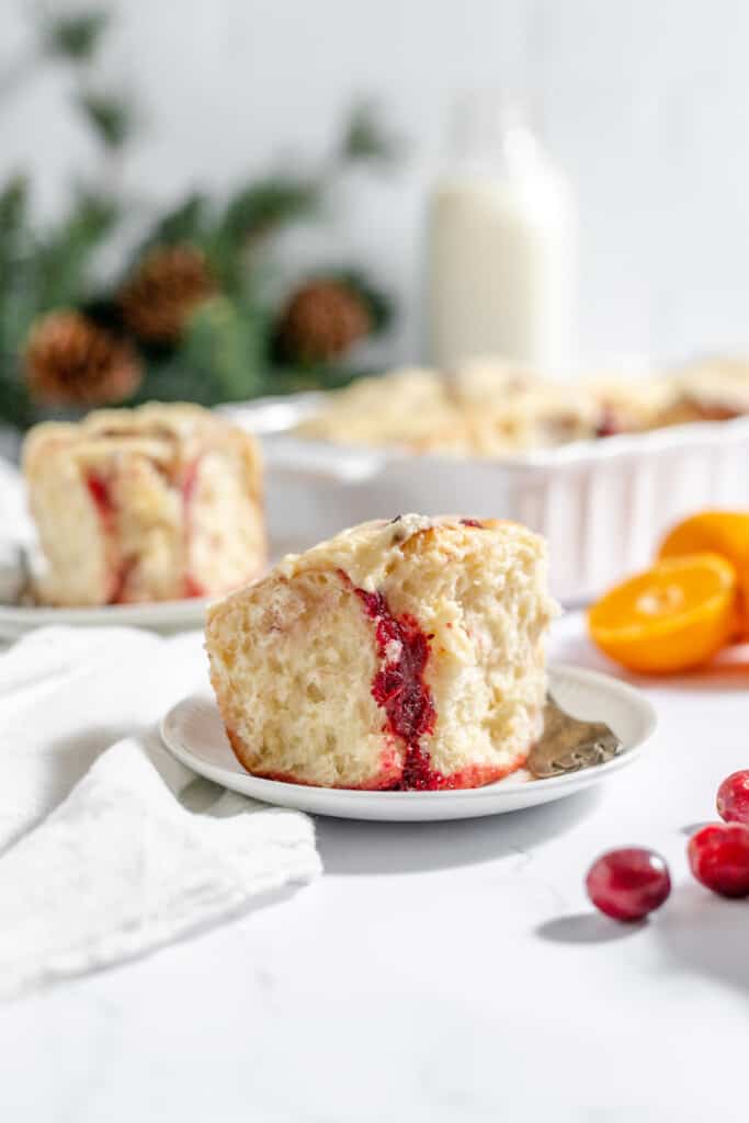 Orange cranberry roll on a plate with cranberries, sliced orange, and a bottle of milk in the photo. 