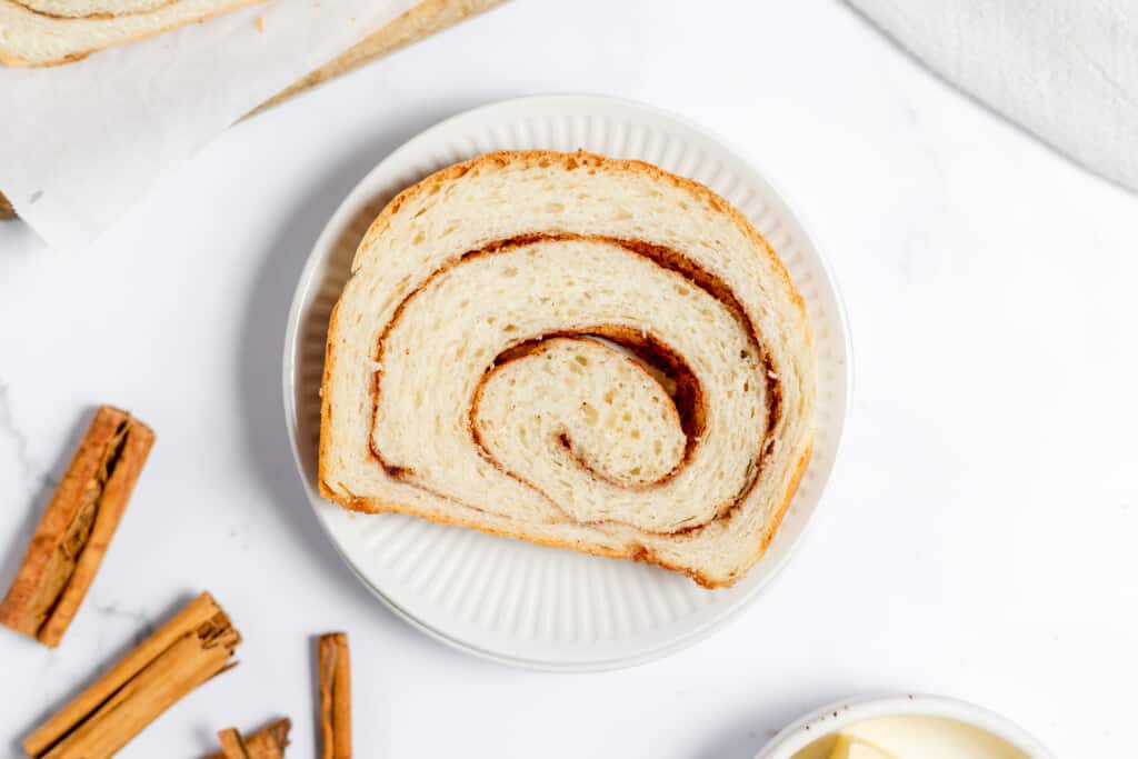 One slice of cinnamon swirl bread on a plate with cinnamon sticks next to it.