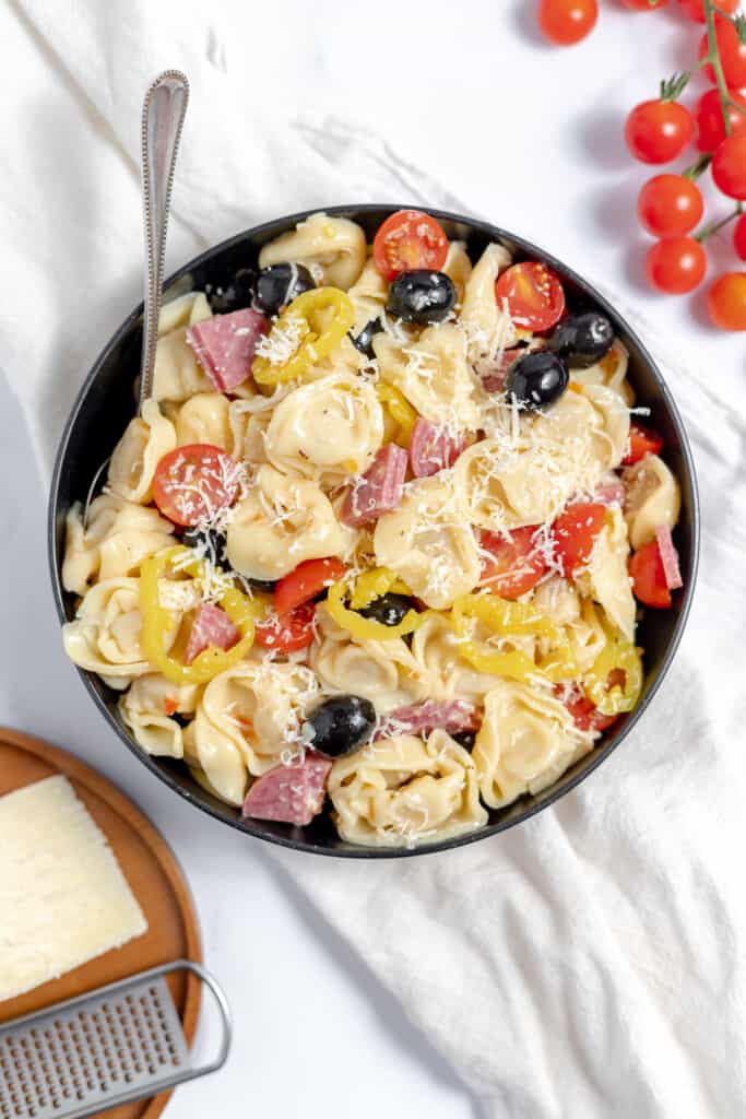 A large bowl of tortellini salad on a tablecloth with parmesan cheese and tomatoes on the side. 