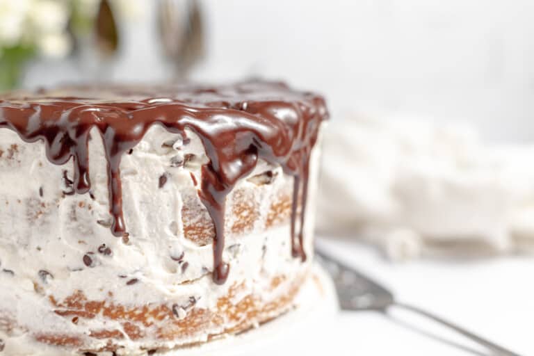 A layered cannoli cake with chocolate ganache dripping down its sides, displayed on a white table with a cake server nearby.