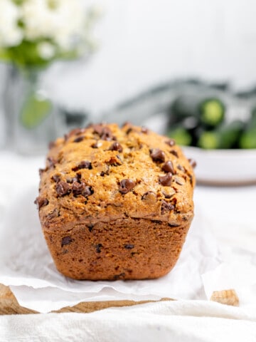 Loaf of chocolate chip zucchini bread on a plate with zucchini piled in the background.